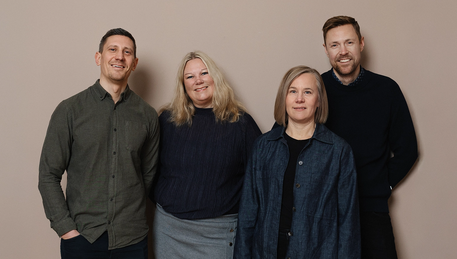 Fyra personer står vid en rosa vägg, ler och tittar in i kameran.